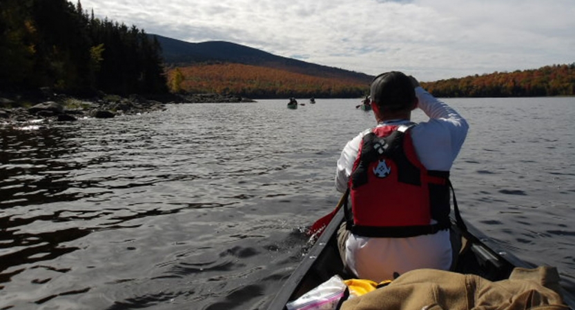 From the back of a canoe, you can see the person in front paddling. The boat is on calm water with trees lining the shore. 